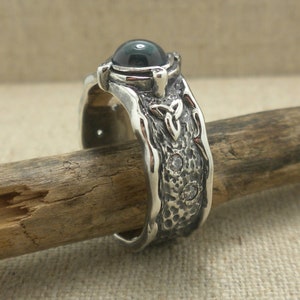May include: A silver ring with a dark green gemstone and a Celtic knot design. The ring is set with small white gemstones.