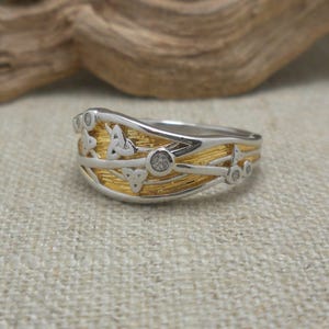 May include: A two-tone ring with a silver band and gold-coloured centre. The ring features Celtic knot designs and small, clear gemstones. The ring is set against a neutral background.