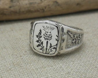 Puede incluir: Un anillo de sello de plata con una detallada grabación de una flor de cardo.