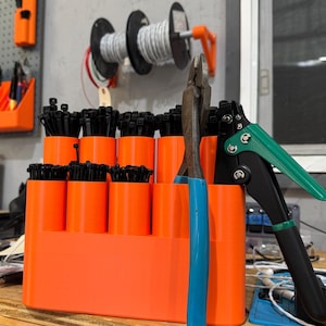 Puede incluir: Un organizador de plástico naranja con 10 compartimentos contiene bridas de cable negras. Un par de alicates de corte de alambre azules y una herramienta de sujeción de bridas de cable verde y negra están al lado del organizador. El organizador está sobre una superficie de madera. El texto "MR. J'S WORKSHOP" está en un tablero de clavijas gris en el fondo.