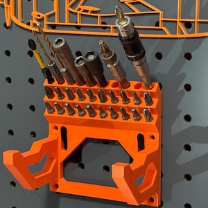 Op de afbeelding: Een oranje 3D-geprinte boorbit organizer gemonteerd op een grijs pegboard. De organizer bevat diverse boorbits en schroevendraaierbits. Een bijpassende oranje omtrek van een voertuig bevindt zich boven de organizer.