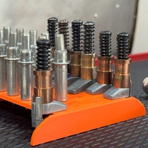 Puede incluir: Un surtido de componentes metálicos y cobrizos, algunos con muelles negros, se disponen sobre una bandeja naranja. Los componentes tienen varias formas y tamaños, lo que sugiere que son piezas para un mecanismo o herramienta más grande.