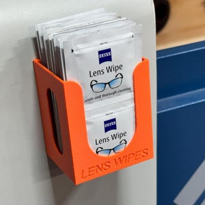 May include: An orange plastic holder mounted on a wall, filled with individual packets of Zeiss Lens Wipes. The packets are white with blue text and a glasses graphic. The holder has the words "LENS WIPES" printed on it.