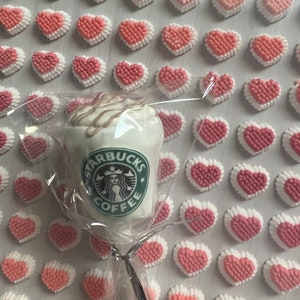 May include: A white chocolate Starbucks coffee cup lollipop with a white and brown swirl on top. The lollipop is surrounded by pink and white heart-shaped candies.