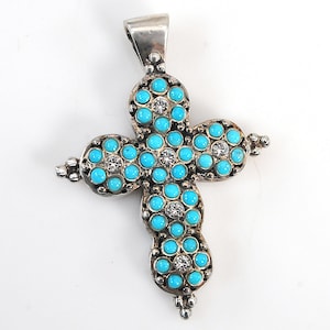 Peut inclure: Un pendentif croix en argent orné d'une série de petites pierres turquoise rondes et vives disposées en grappes. Chaque grappe est centrée par une pierre blanche transparente et facettée. Le métal présente une finition oxydée sombre.