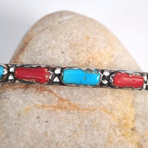 Puede incluir: Una pulsera de plata con piedras rectangulares turquesas y rojas alternas. La pulsera se presenta sobre un fondo neutro, resaltando los colores vibrantes de las piedras. La pulsera es una joya.