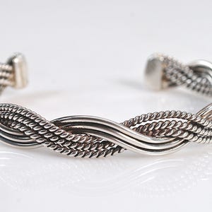 May include: A silver cuff bracelet featuring a multi-strand design with alternating smooth polished wires and twisted rope-textured wires woven together in a continuous braided pattern, ending in two solid, rounded silver terminal caps.
