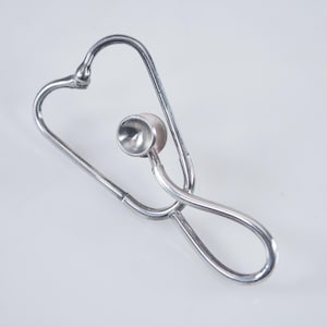 May include: A silver-toned metal brooch shaped like a medical stethoscope. The design features a continuous wire frame forming the binaurals and tubing, with a small, circular chest piece attached to the center of the structure.