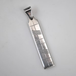 以下が含まれることがあります： 長方形の形をしたシルバーペンダント。上部にループが付いています。ペンダントには、一連の線と正方形がエッチングされており、幾何学模様を作り出しています。シンプルでありながらエレガントなジュエリーです。