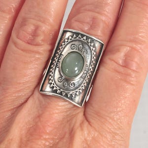 May include: A rectangular silver ring featuring a central oval-shaped, smooth, pale green stone. The silver band has a wide, curved face with intricate embossed detailing surrounding the stone, including a scalloped border and small scroll motifs.