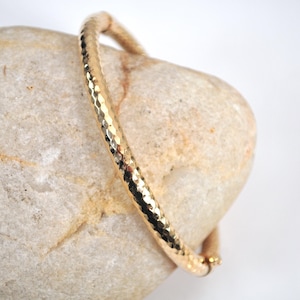 May include: A gold-colored bangle bracelet with a hammered texture. The bracelet is resting on a light-colored stone. The bracelet has a simple, elegant design and a secure clasp. The bangle is approximately 2.5 inches in diameter.