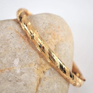 May include: A gold-colored bangle bracelet with a textured, diamond-patterned design. The bracelet is resting on a light-colored stone, showcasing its intricate details and metallic sheen. This piece of jewelry is a classic accessory.