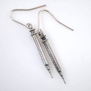 May include: A pair of silver-toned syringe-shaped earrings. Each earring features a long, slender barrel with etched details and a pointed tip. The earrings have a hook closure.
