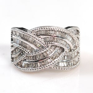 Puede incluir: Un anillo ancho de tono plateado con un diseño entrelazado complejo. La superficie está densamente engastada con numerosas piedras transparentes pequeñas de corte baguette rectangular. Los bordes tienen un borde metálico trenzado y texturizado.