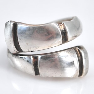 Peut inclure: Deux bagues argentées entrelacées, présentant un design moderne. Chaque bague a une forme rectangulaire avec un détail de ligne sombre en retrait. Les bagues sont posées sur un fond blanc uni, soulignant leur éclat métallique.