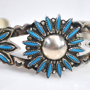 Könnte beinhalten: Ein silbernes Armreif-Armband mit einem zentralen Sonnenstrahl-Design. Der Sonnenstrahl hat eine große silberne Mitte, umgeben von türkisfarbenen, tropfenförmigen Steinen. Das Armband hat dekorative Blatt- und Blumenakzente mit weiteren türkisfarbenen Steinen.