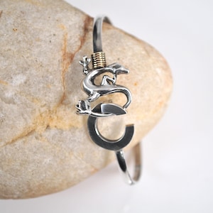 Könnte beinhalten: Silberfarbenes Metallarmband mit einem Eidechsen-Design. Die Eidechse sitzt auf einem gebogenen, buchstabenförmigen Element. Das Armband hat ein schlichtes, elegantes Design, das für den Alltag geeignet ist. Die Eidechse hat aufwendige Details.