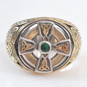 May include: A two-tone ring featuring a silver and gold Celtic cross design. The center of the cross is set with a small, round, dark green stone. The band and the outer edges of the cross are decorated with intricate, embossed knotwork patterns.