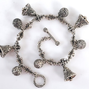 May include: A silver-toned charm bracelet featuring alternating bell-shaped and spherical charms with intricate swirling patterns. The bracelet includes a toggle clasp with the word INDONESIA engraved on the bar and a circular ring closure.
