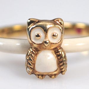May include: A gold-toned owl ring with white enamel details. The owl is the focal point, perched on a cream-colored band. The ring has a polished finish, suitable for everyday wear.