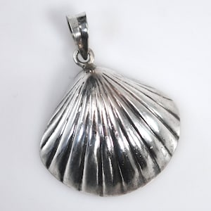 May include: A silver pendant in the shape of a scallop shell. The pendant has a loop at the top for a chain and features detailed ridged lines radiating from the center. The shell design is highly polished, reflecting light.