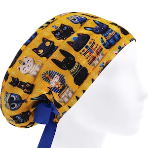 May include: A yellow surgical scrub cap with a repeating pattern of black cat illustrations, some with Egyptian motifs. The cap, with a blue tie, is worn by a mannequin head. The cat designs are in different poses and styles, adding visual interest.