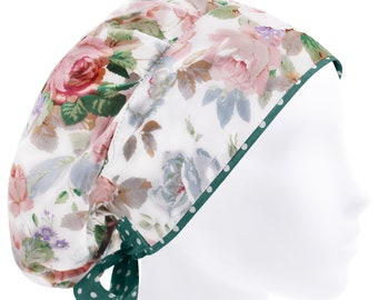 May include: A scrub cap featuring a white background with a floral pattern of pink roses, green leaves, and small purple flowers. The cap has a dark green trim with white polka dots and matching green polka dot ties at the back for adjustment.
