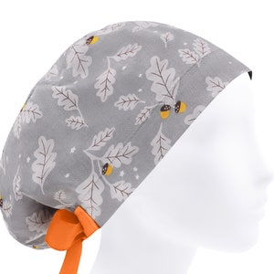 Puede incluir:  gorro de quirófano gris con un patrón de hojas de roble y bellotas. El gorro tiene una corbata naranja brillante.