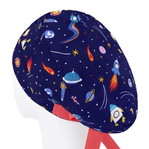 K&ouml;nnte beinhalten: Eine dunkelblaue OP-Haube mit einem bunten Weltraummuster aus Raketen, Planeten, Sternen, Kometen und fliegenden Untertassen. Die Haube verf&uuml;gt &uuml;ber leuchtend korallenfarbene Stoffb&auml;nder an der Basis, die f&uuml;r einen sicheren Sitz w&auml;hrend der Arbeit sorgen.