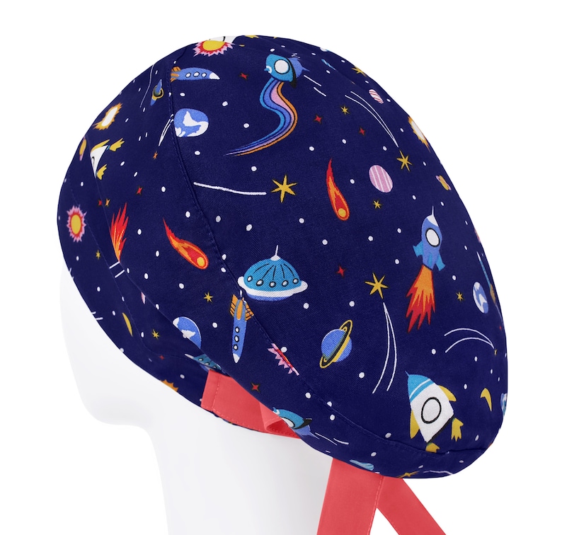 K&ouml;nnte beinhalten: Eine dunkelblaue OP-Haube mit einem bunten Weltraummuster aus Raketen, Planeten, Sternen, Kometen und fliegenden Untertassen. Die Haube verf&uuml;gt &uuml;ber leuchtend korallenfarbene Stoffb&auml;nder an der Basis, die f&uuml;r einen sicheren Sitz w&auml;hrend der Arbeit sorgen.