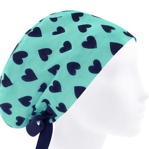 May include: A turquoise blue scrub hat with a navy blue heart pattern. The hat has a tie at the back.