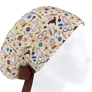 May include: A cream-coloured surgical scrub cap with a repeating pattern of owls, spectacles, letters, and other magical-themed illustrations. The cap has a brown bow at the back. The design is reminiscent of a popular fantasy book series.