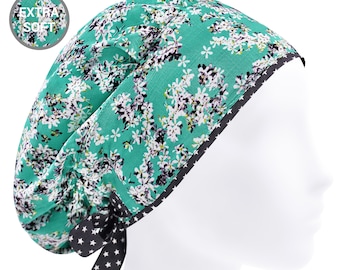 May include: A teal scrub cap featuring a white and black floral pattern with a black trim decorated with white stars. The cap includes a tie-back closure with matching star-patterned ribbons and a circular badge in the corner reading EXTRA SOFT.