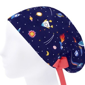 K&ouml;nnte beinhalten: Eine dunkelblaue OP-Haube mit einem bunten Weltraum-Muster aus Raketen, Planeten, Sternen und Kometen. Die Haube verf&uuml;gt &uuml;ber ein einfarbiges, korallenrotes Band am Hinterkopf, mit dem die Passform individuell angepasst werden kann.
