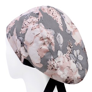 May include: A grey and pink floral print scrub hat with a black tie. The hat has a floral pattern with pink flowers and leaves on a grey background.