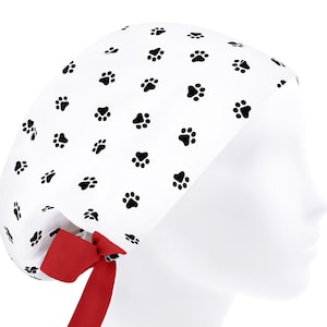 Op de afbeelding: Witte chirurgische scrub-muts met een zwart pootprintpatroon en een rode strik.