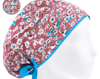 May include: A scrub cap featuring a dusty rose background with a white and bright blue floral pattern. The cap has a bright blue trim and matching blue ribbon ties at the back. A circular inset in the top left corner displays the text EXTRA SOFT.