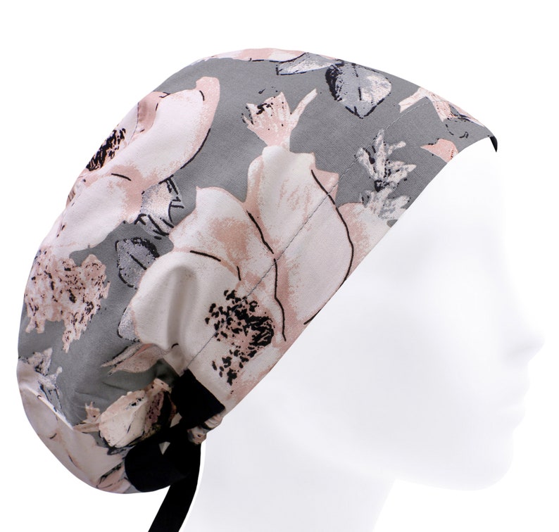 May include: A grey and pink floral scrub cap with black ties. The cap has a large floral print with pink and white flowers on a grey background.