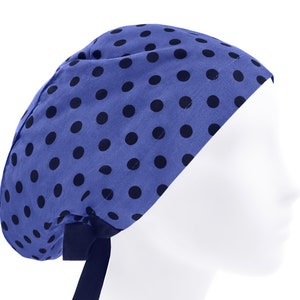 Può includere: Una cuffia chirurgica blu con un motivo a piccoli pois neri regolarmente distanziati. La cuffia presenta una calotta arrotondata e include un laccio in tessuto nero sul retro, annodato con due estremità libere che pendono dalla base.