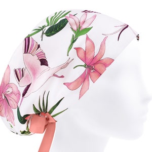 May include: A white scrub cap featuring a botanical print with pink lilies, green tropical leaves, and flying cranes. The cap includes a solid coral-colored ribbon tie at the back, designed to secure the fabric around the head for a comfortable fit.