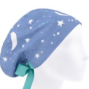 Könnte beinhalten: Eine blaue OP-Haube mit einem Muster aus weißen Mondsicheln und Sternen. Die Haube hat einen gerafften Hinterkopfbereich mit einem türkisfarbenen Bindeband. Der Stoff zeigt einen Himmelsdruck mit weißen Sternen in verschiedenen Größen und Punkten.