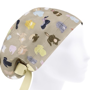 Op de afbeelding: Een beige scrubmuts met een patroon van cartoon konijnen in verschillende kleuren en poses. De muts heeft een gele strik.