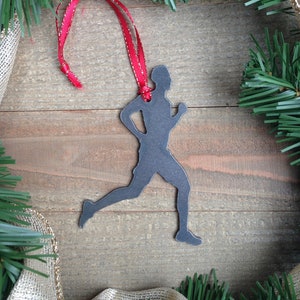 May include: A black metal silhouette of a runner ornament with a red ribbon. The ornament is on a wood background with green pine branches.