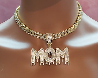 May include: Gold chain necklace with a large, diamond-encrusted pendant that says "MOM" in dripping letters.