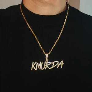 Könnte beinhalten: Eine goldene Kettenkette mit einem Anhänger, auf dem in einer stilisierten Schriftart mit Diamanten "KMURDA" steht.