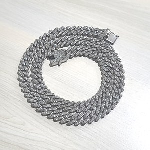 Puede incluir: Un collar de cadena de plata con un diseño de eslabón cubano. La cadena está cubierta de pequeñas piedras transparentes que le dan un aspecto brillante.