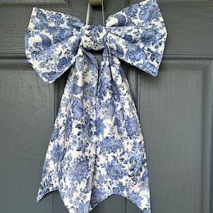 May include: A large, decorative bow made of fabric with a blue floral pattern on a white background. The bow is hanging on a door. The bow's design features a classic floral print, adding a touch of elegance to any space.