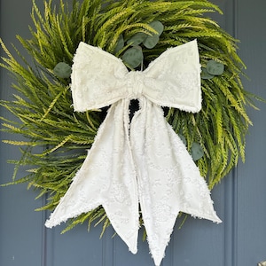May include: A decorative wreath featuring a vibrant green base with faux foliage and eucalyptus accents. A large, textured white bow is centered on the wreath, adding a touch of elegance. The wreath is displayed on a blue door.