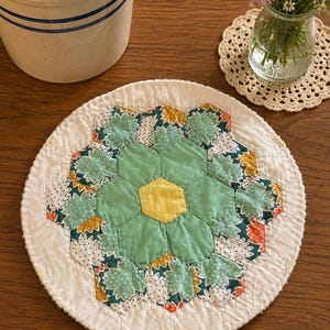 May include: A round quilted fabric mat featuring a central yellow hexagon surrounded by light green, teal, and white floral patterned hexagons. The piece is finished with a white border and visible stitching, creating a flower-like geometric design.