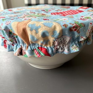 May include: A light blue bowl cover with a festive Christmas pattern featuring gingerbread men, candy canes, and cookies. The cover has a ruffled edge and is placed over a white bowl. The background is a gray surface.